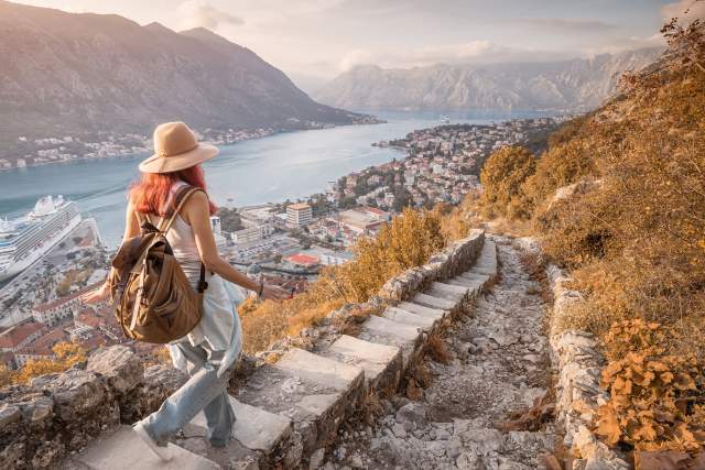 A woman with a backpack walks down steps by a lake with scenic mountains in the distance.