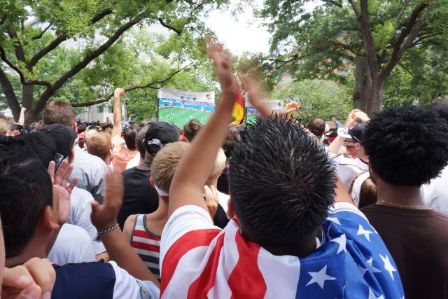 A group of fans dressed in USA apparel watch a soccer match on a big screen while cheering on.