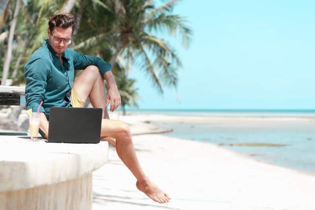 A man sits on a ledge near the beach while working on his laptop.