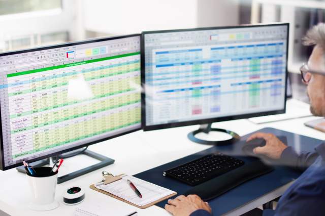 A person using double monitors to browse through several Excel spreadsheets.