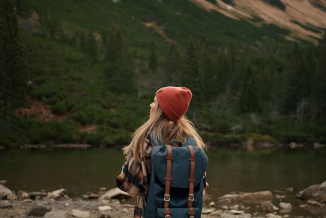 A woman in hiking gear stands by a lake in the middle of the mountains.