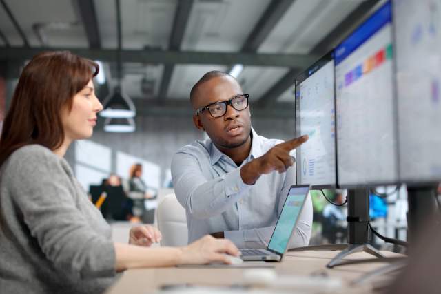 Two professionals review CRM dashboard data on dual monitors during a meeting sales strategy session, illustrating practical CRM tactics and best practices for improving sales team performance and adoption.