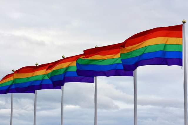 Six Pride flags blowing in the wind with cloudy skies.