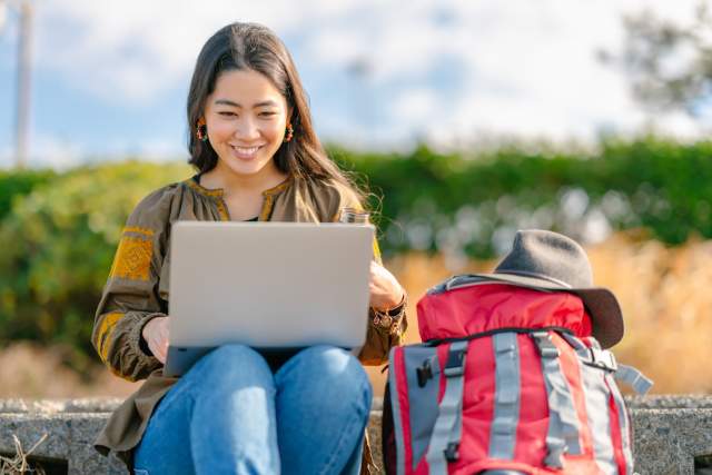 A traveller sits outdoors with a laptop and backpack, using digital tools to plan their trip.