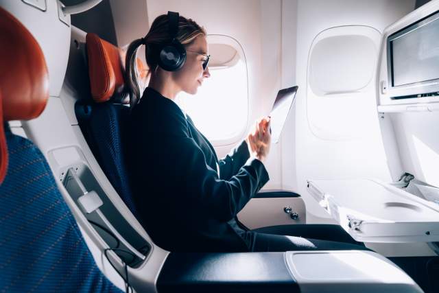 A woman travels in an airplane with headphones on while viewing a tablet, representing technology in travel and tourism.