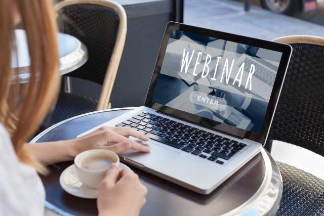 A person seated at a small table with a cup of coffee getting ready to watch a webinar on their laptop.