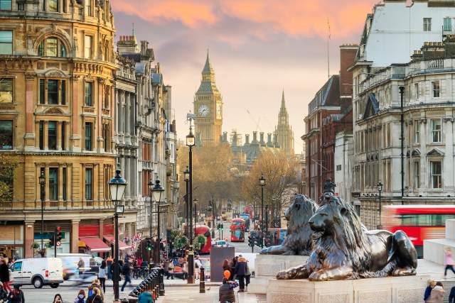 View of Big Ben and Westminster from Trafalgar Square in London at sunset, featuring historic architecture, lion statues, and red double-decker buses.