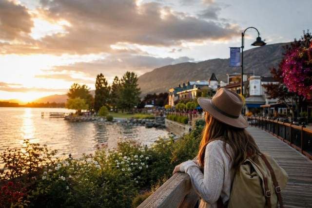 Traveler with a backpack overlooks a lakeside boardwalk at sunset, with mountains, waterfront buildings, and colorful flowers.