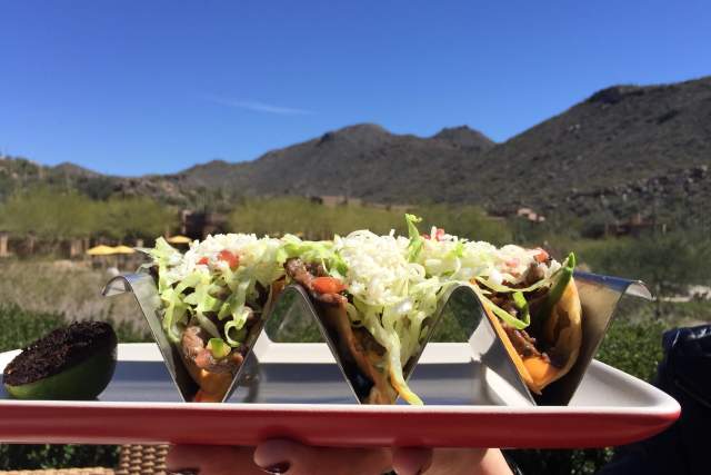A person holds a plate of three tacos with Sonoran Desert foothills in the background.
