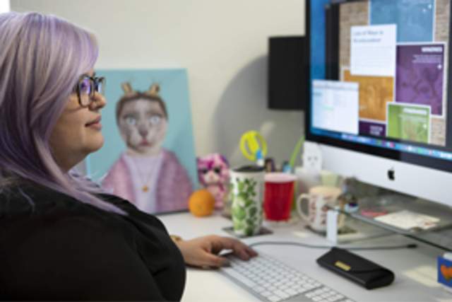 A woman with lavender hair and glasses designs a website on a desktop.