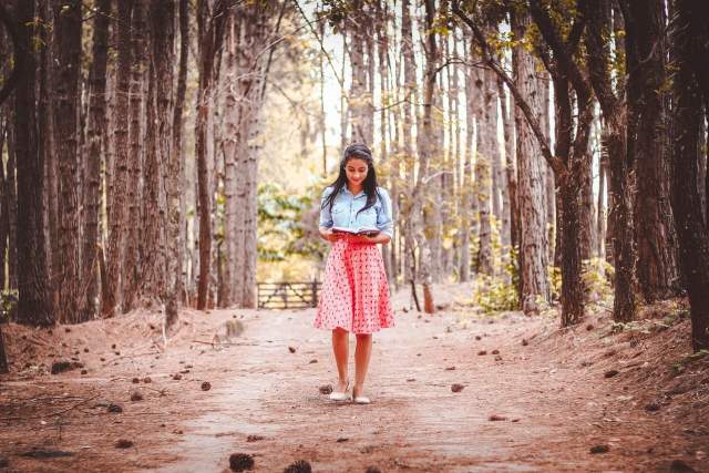 A woman walks down a path through the woods while reading a book.