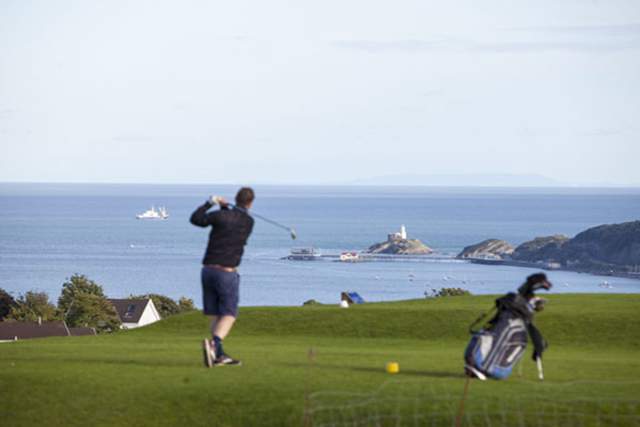 Golfer swinging club with Mumbles lighthouse and sea views in Swansea Bay.