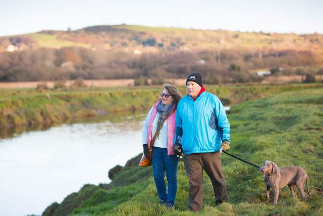 A man and a woman walking on a river bank with a brown dog. The dog has a stick in its mouth.