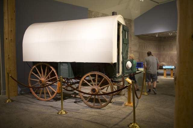 A restored covered wagon on display behind ropes inside the Buffalo Bill Center of the West, surrounded by museum visitors.