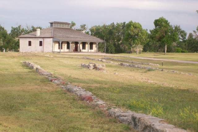 Historic stone foundations leading to a preserved 19th-century military building surrounded by trees and open prairie.