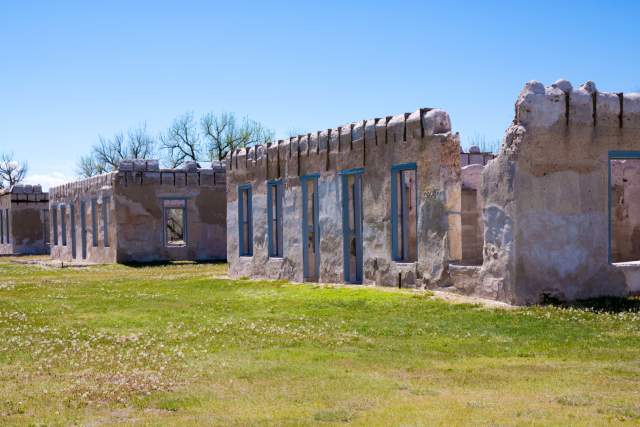 Crumbling adobe and stone ruins of historic Fort Laramie under a clear blue sky with scattered trees and green grass.