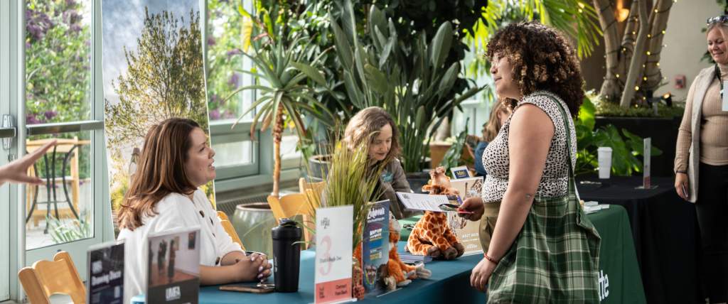 In a greenhouse room with lots of plants a woman is standing in front of colorful vendor tables talking to the people sitting behind them.