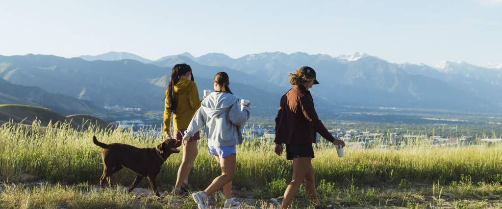 Three girls walking with a dog on a mountain trail between green grass with the Salt Lake Valley and Wasatch mountains in the background