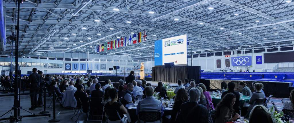 Speaker on stage at the Utah Olympic Oval, people sitting at round tables in the foreground, UT Hockey Club and Olympic logos in the background