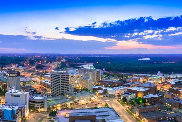 Panoramic sunset view of downtown Montgomery skyline with city lights, riverfront buildings, and Alabama horizon at dusk glow.