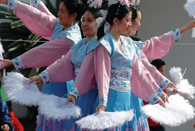 4 women of Asian descent dressed in native costumes in light blue and pink, holding white feather fans