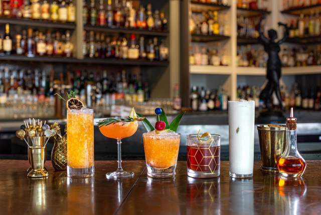 Assorted craft cocktails arranged on a wooden bar counter with shelves of liquor bottles in the background at a Houston bar.