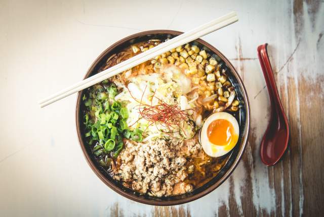 Bowl de ramen servido en un tazón grande con huevo, maíz, cebollín, carne molida y fideos, acompañado de palillos sobre una mesa de madera.