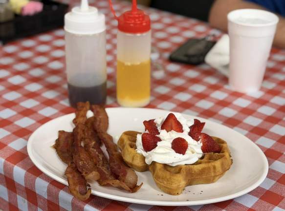 Strawberry and whipped cream over a Texas Shaped waffle next to 4 strips of bacon