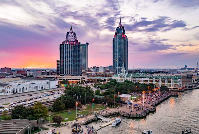 A vibrant sunset over Downtown Mobile skyline, featuring tall buildings, a bustling waterfront, and a lively crowd below.