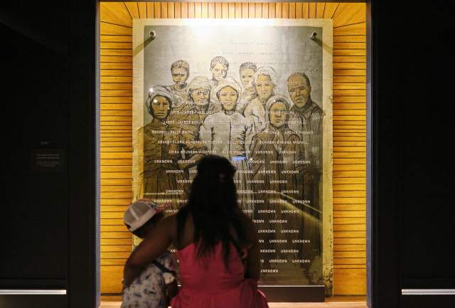 A Black woman with long hair holds her child while they look at a photo collage of formerly enslaved residents of Africatown, Alabama