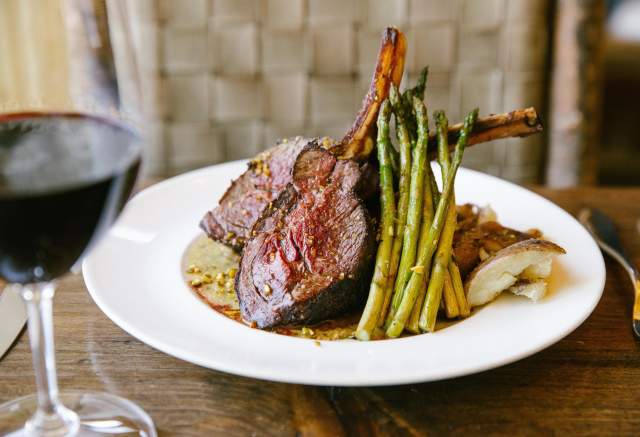 A plate featuring two lamb chops, asparagus, and a side of mashed potatoes, accompanied by a glass of red wine. Elegant dining setting.