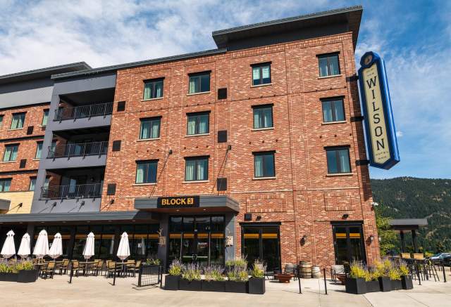 Exterior view of a modern brick hotel with a sign reading "WILSON." Outdoor seating area with umbrellas and planters in front. Scenic mountains in the background.