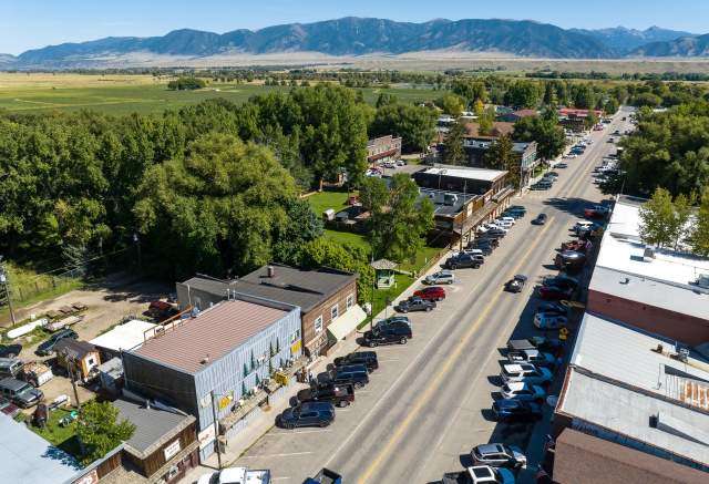 Aerial view of a charming small town street lined with shops and parked cars, surrounded by lush greenery and distant mountains.