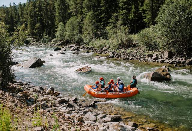 A bright orange raft with six people navigates a rushing river, surrounded by lush green trees and rocky banks. The scene captures an adventurous outdoor experience.