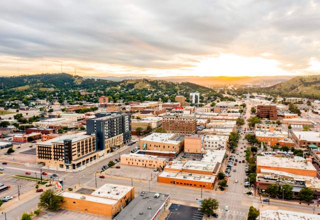 Aerial view of downtown Rapid City at sunset, with downtown streets lined by low-rise buildings, cars, and parking lots. Rolling green hills surround the city, while dramatic clouds stretch across the sky and golden sunlight glows on the horizon.