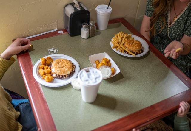 Two women sit at a table with cheeseburger meals sitting in front of them. One holds a French fry in her hand. Two Styrofoam drink cups sit beside their plates.