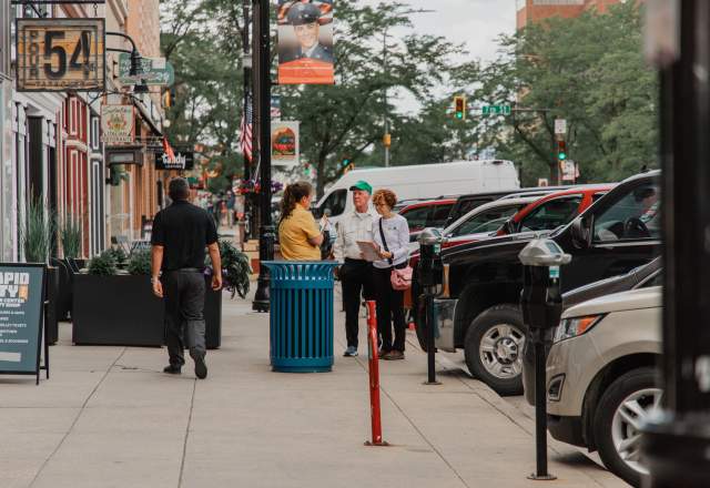 Downtown Rapid City Ambassador in a yellow shirt speaks with visitors on a busy sidewalk lined with storefronts, signs, parked cars, and street banners.