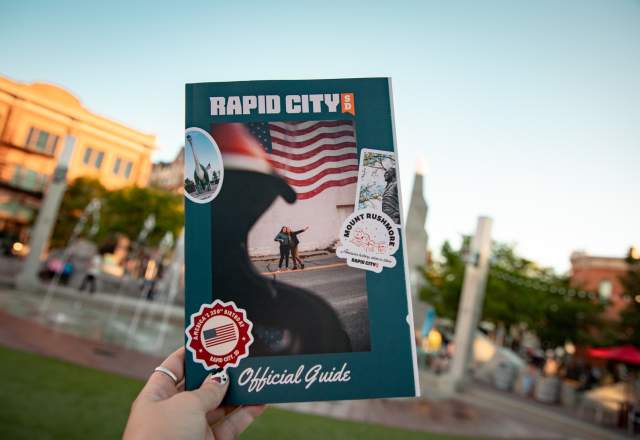 The cover of the Visit Rapid City guide is held up by a woman against the backdrop of the fountains at Main Street Square.