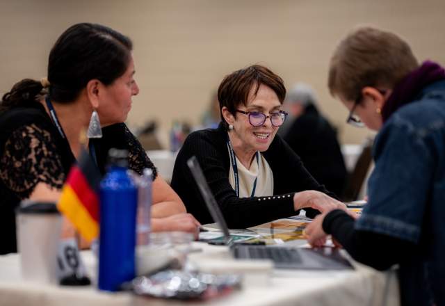 Three people engaging in a lively discussion at a table with laptops and papers. A small German flag and a blue water bottle are visible. The mood is collaborative.