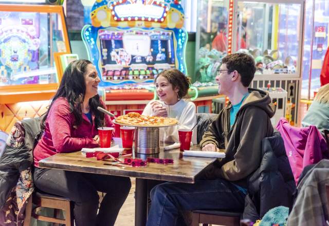 Family of three sitting around a table eating pizza with arcade games in the background at Press Start in Rapid City, SD