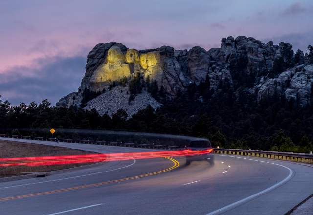 Mount Rushmore, lit up by yellow floodlights at dusk, sits in the background as a vehicle traveling down the highway is trailed by a long line of red taillights, created by a long-exposure photo.