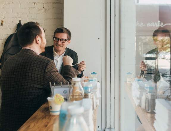 Two men, both wearing dark suits, dine at a windowside counter. They are engaged in conversation.