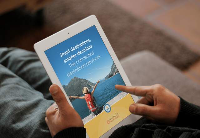 Person holding a tablet displaying a travel playbook cover titled “Smart destinations, smarter decisions: The connected destination playbook” with a lakeside mountain scene.