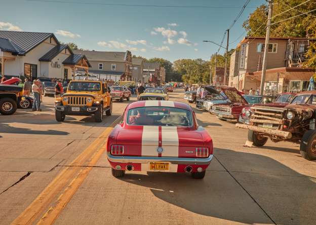 A classic red car driving down a street lined with other classic cars.