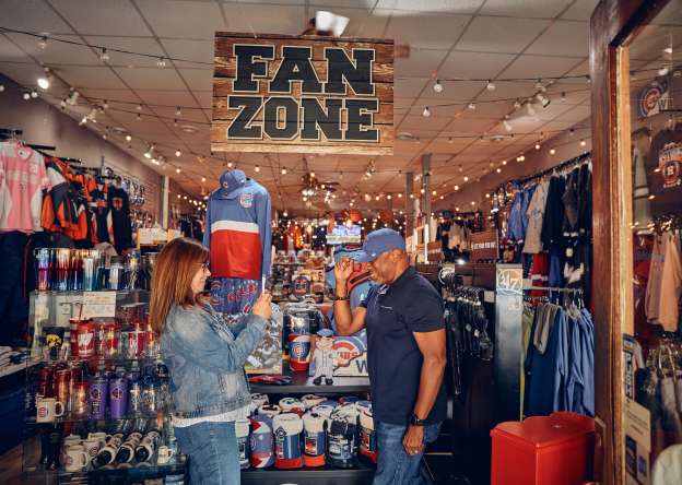 Two people in a sports store, one person taking a picture of the other posing with a hat. A sign above them says, "Fan Zone".