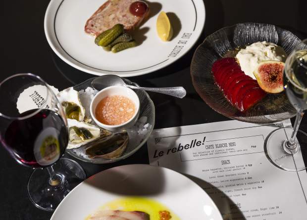 3 plates of food, 2 wine glasses and a Le Rebelle menu on a dark table