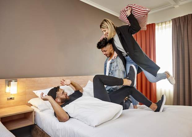 Three friends having a pillow fight in one of Ibis Perth's hotel rooms.