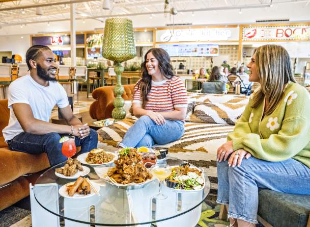 Three people enjoy expertly crafted food and beverages with smiles, gathered at a table in the brightly lit and beautifully decorated Politan Row food hall in Peachtree Corners.