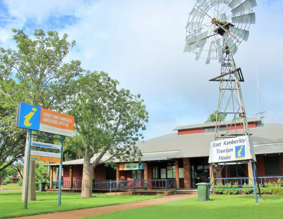 Image depicts the Kununurra Visitor Centre from a front, slightly off centre angle. In the foreground stands an old windmill with the sign on and some boab trees and a standing sign on the opposite side of the footpath. The building is brick with a tin roof.