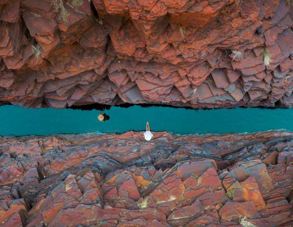Overhead shot of two people in a narrow section of Weano Gorge, Karijini National Park. One is walking along the gorge floor, through water, and one is relaxing on the rocks.